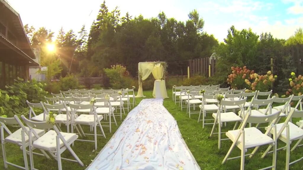 Allée fleurie menant à une arche de cérémonie en extérieur, décorée de voilage et de chaises blanches disposées dans un jardin verdoyant au coucher du soleil – Agence Moment de Grâce.