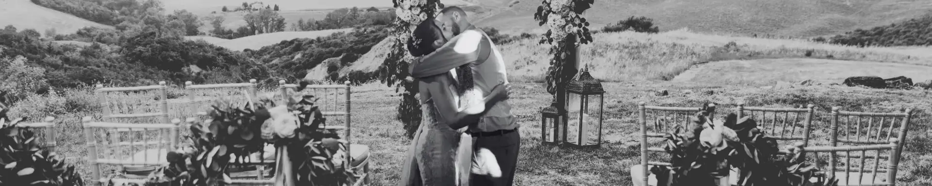 Cérémonie laïque en extérieur organisée par l’Agence Moment de Grâce, avec un couple s’enlaçant devant une arche fleurie, des chaises décorées et un panorama de collines verdoyantes.