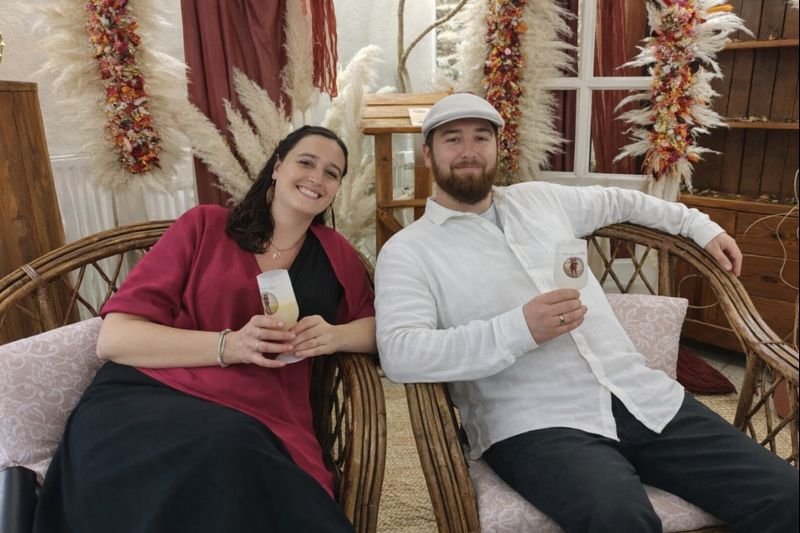 Deux personnes assises dans un fauteuil en rotin, souriantes, tenant des verres personnalisés dans une décoration bohème avec pampas et guirlandes florales.